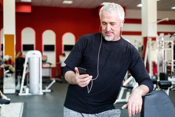 Gray-haired man athlete stands in the gym listening to music through headphones holding the phone