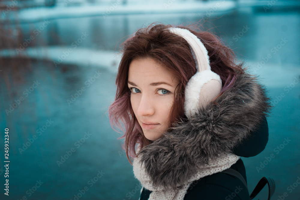 Wall mural girl with red hair in a dark winter jacket and hood with fur in fur pink headphones in the park against the backdrop of the lake