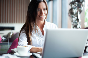 Young smiling businesswoman on a break in a cafe. She is working at laptop and drinking coffee