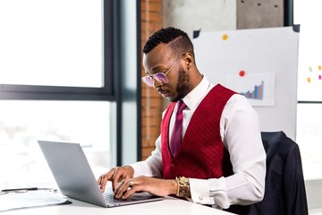Fototapeta premium Stylish African businessman in a suit working at a laptop at a table in a modern office