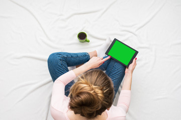 Woman woman uses a tablet, sitting on a white crumpled blanket, green Cup of coffee or tea, women's feet. Women's hands. Background with copy space, for advertisement. Top view