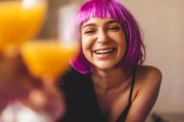 Attractive playfull woman wears pink wig, black lingerie and shirt. Girl drinking orange juice in glass. Woman sitting on the floor. She look sexy and hot, girl raised a glass. Toast