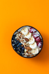 Healthy breakfast berry smoothie coconut bowl topped with blueberry,almond, banana, granola and raspberries. Top view. orange background.