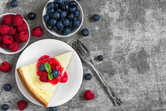slice of new york cheesecake with fresh raspberries, blueberries, jam and mint with fork on concrete background