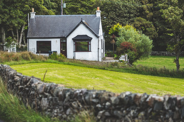 Fototapeta na wymiar Small white countryside cottage surrounded by the garden. Islay island. Scotland. Green grass covered lawn before the cozy farm house. Ideal background for the fairy tale collages and illustrations.
