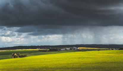 Obraz premium Grey rainy sky above the green meadow. Northern Ireland landscape. Dense dark clouds under the grassland and village. Stunning countryside Irish scenery. Horizon panoramic view. Contrast colors.