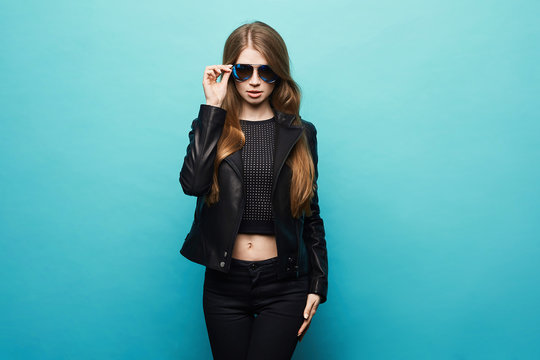 Fashionable blonde model girl with perfect body in the black leather jacket and in stylish sunglasses posing at the blue background