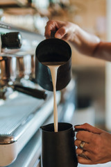 Barista making cafe latte in coffee shop, female hands pouring milk into a black pitchr to steam milk and make cappuccino or latte in a coffee shop