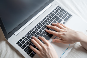 Close-up of woman's hand using laptop with blank screen on bed in home interior. The light from the screen illuminates the female hands on the laptop keyboard.