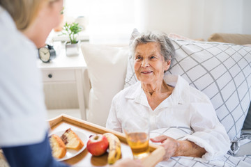 A health visitor bringing breakfast to a sick senior woman lying in bed at home.