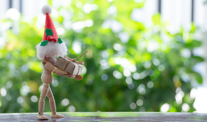 A man model made of wooden stand holding a gift box  and wearing a Santa's hat. The background is a bokeh  from a tree. Concept send gifts on special days, Christmas,New Year