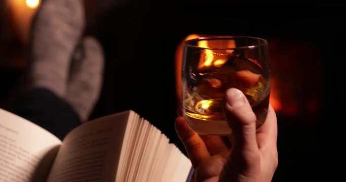 Man Reading Book And Drinking Glass Of Whisky Warming Feet By Flames Of Wood Burning Stove 