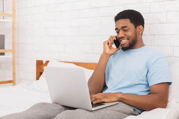 Smiling man talking on phone and using laptop in bed