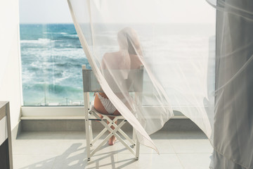 Naked beautiful female in the bikini sitting on a chair behind the transparent waving curtains on the glass balcony at the summer windy day. Sea view