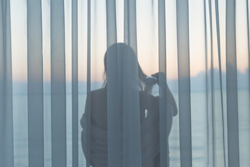 Naked beautiful girl stands behind the transparent curtain combing her hair on the balcony early morning at sunrise. Sea view.