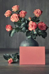Bouquet of pink roses and a blank pink paper card on dark rustic background, text space