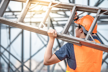 Young Asian maintenance worker man with orange safety helmet and vest carrying aluminium step...