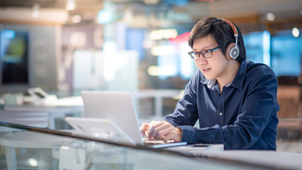 Asian business man listening to music by headphones while working with laptop computer in coworking space. Freelance or digital nomad lifestyle. Online shopping or E-commerce social media on internet