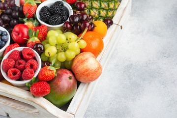 Healthy fruits in wooden tray, strawberries raspberries oranges plums apples kiwis grapes blueberries mango persimmon pineapple on the white table, copy space for text, selective focus