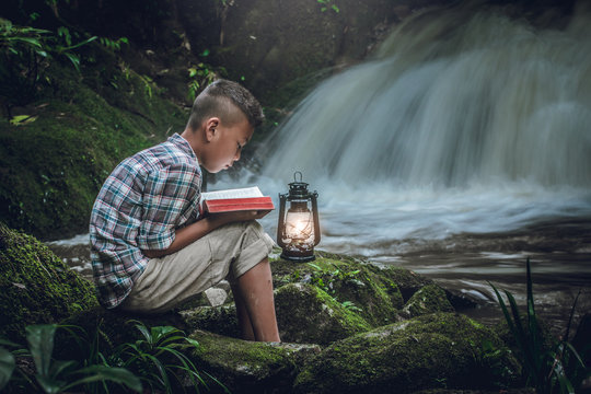 Boy siting and reading book or bible on green rock with oil lamp at waterfall. Children and religion.