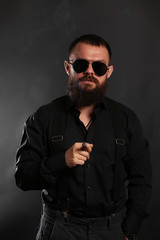 young handsome man with big beard and black sunglasses on black background  smoking cigar 