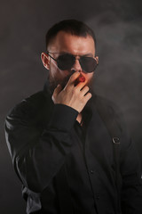 Obraz premium young handsome man with big beard and black sunglasses on black background smoking cigar 