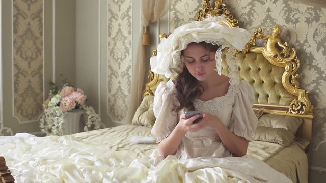Cute young woman in ball gown sitting on the gold decorated bed and texts on cell phone using gadget