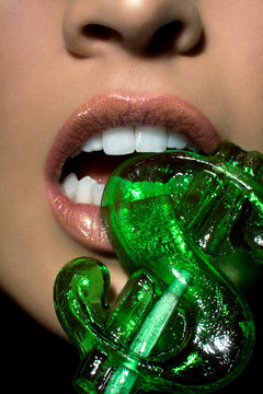 Close up of a young woman's lips wearing flesh colored lipstick and a dollar sign lollipop between her teeth.