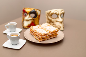 Typical Italian carnival fritters (Chiacchiere di Carnevale) dusted with powdered. Composition with two cups of coffee and in the background are Venetian carnival masks. Italian Carnival sweets. 