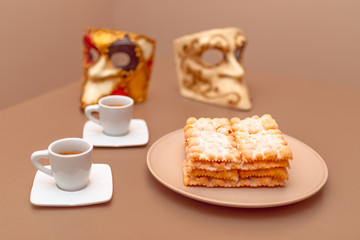 Typical Italian carnival fritters (Chiacchiere di Carnevale) dusted with powdered. Composition with two cups of coffee and in the background are Venetian carnival masks.