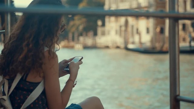 Young woman using smartphone in street of Venice with the channel and boat in background. Woman holding a cell phone and touch screen for surfing internet and checks social network