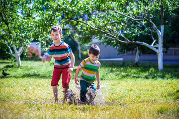 Fototapeta premium Little boy and his brother play in summer park. Children with colorful clothes jump in puddle and mud in the garden.