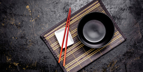 Empty Japanese dishes. A black ceramic bowl for Chinese noodles or Thai soup lies on a bamkuk rug. White saucepot for soy sauce and red Chinese sticks on a black background. Top view, copy space