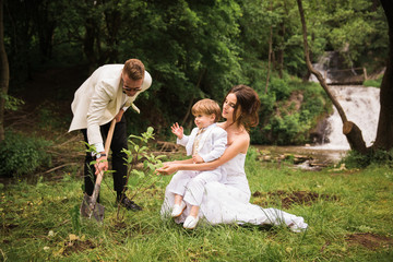 The happy newlyweds planting a tree near the waterfall and a little boy sitting on the lap of a bride and helping them