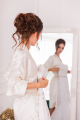 The beautiful young bride standing in front of the mirror indoor and holding her bridal gown
