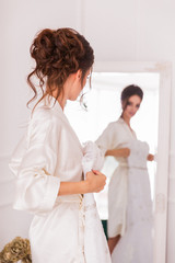 Obraz premium The beautiful young bride standing in front of the mirror indoor and holding her bridal gown