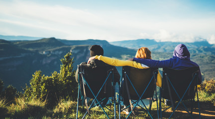 three friends sit in camping chairs on top of a mountain, travelers enjoy nature and cuddle,...