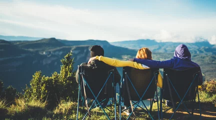Fotobehang drie vrienden zitten in kampeerstoelen bovenop een berg, reizigers genieten van de natuur en knuffelen, toeristen kijken in de verte op de achtergrond van een panoramisch landschap, weekendconcept mockup © A_B_C