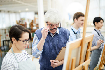Portrait of mature art teacher criticizing work of student painting picture on easel in art class,...