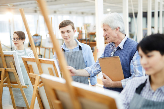 Portrait of mature art teacher helping group of students painting picture on easels in art class, copy space
