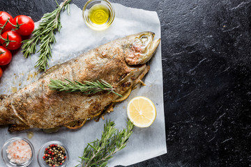 Baked trout fish with salt, lemon and rosemary on paper over black background