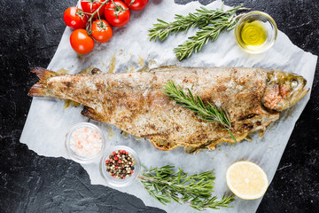 Baked trout fish with salt, lemon and rosemary on paper over black background