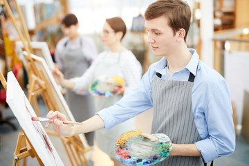 Side view portrait of smiling teenage boy holding palette and painting picture on easel enjoying work in art studio