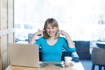 Dont want to hear! Portrait of attractive interesting young girl freelancer in blue blouse are sitting in cafe and making video call on laptop, covering ears with fingers that not hear to companion.