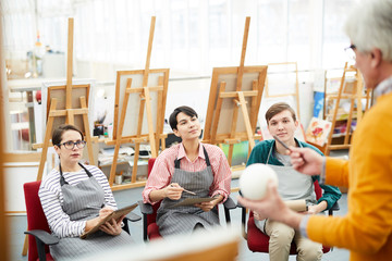 Portrait of group of cheerful art students sitting in row and listening to teacher during sketching session or lecture in class