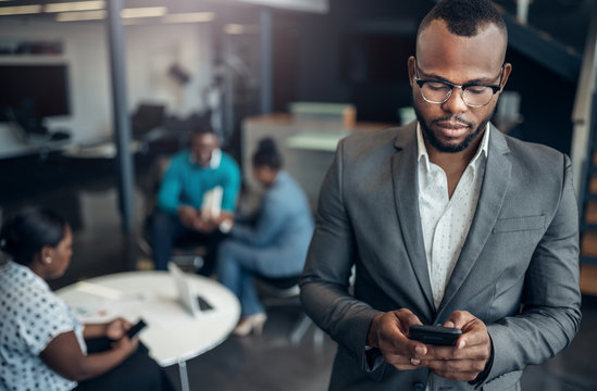 Portrait Of A Confident Black Businessman Looking Down On His Mobile Phone And All African Team In The Background