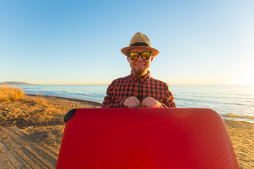 Fototapeta premium Travelling, holidays and people concept - happy man in sunglasses and hat carring his red suitcase near the sea