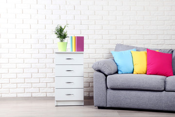 White bedside table near grey sofa and colorful pillows on brick wall background