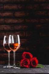 Two wine glasses of rose wine on brick background, bouquet of red roses for romantic evening for Valentines day surprise, marriage proposal passion and love celebration, copy space 