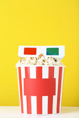 Popcorn in striped bucket with glasses on yellow background